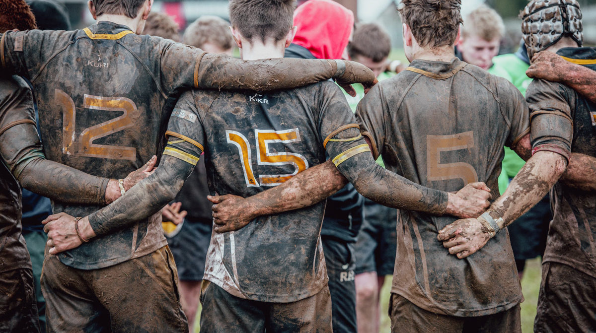 three boys in muddy rugby kits embrace following a schools cup match